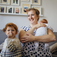 Mother with her two children in the living room of their home. The mother is holding a baby and the other little girl is laughing and looking at the camera while sucking her thumb.