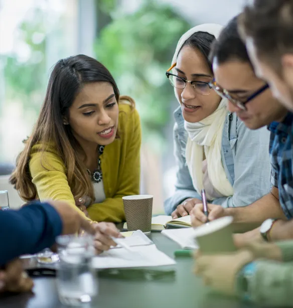 A multi-ethnic group of business people are indoors in an office boardroom. They are sitting around a table and making plans.