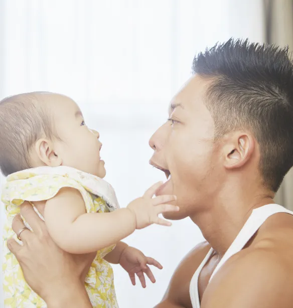 An adult holding a baby, with both facing each other indoors. The adult has their mouth open wide, and the baby is reaching toward their face.