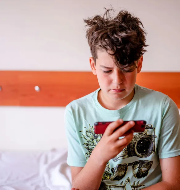 A young boy sits in his bedroom playing on a mobile device.