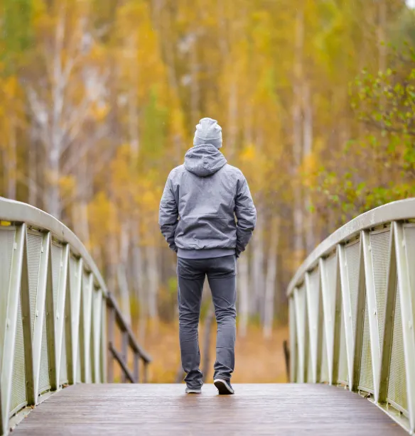 Young person in grey clothes slowly walking on bridge at natural park in colourful autumn day. Spending time alone in nature.