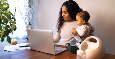 A young mixed race African American mother holds her daughter while taking notes at her dining table serving as a temporary remote work from home station with breast pump in foreground.