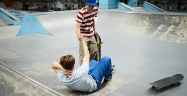 Two teenage boys are in a skate park. One boy is laying on the ground on his back, he has fallen. The other has extended his arm to help him back on his feet. 