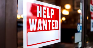Sign text closeup for help wanted with red and white colors by entrance to store shop business building during corona virus covid 19 pandemic. Sign text closeup for help wanted with red and white colors by entrance to store shop business building during corona virus covid 19 pandemic.