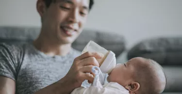 Young father feeding his baby boy son with milk bottle at living room during weekend