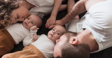 Happy young family with newborn twin babies lying on the bed together, parents holding hands and kissing their babies