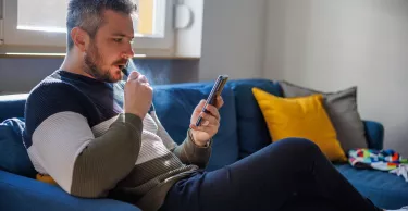 Man sitting on the sofa at home and using smart phone for home finances, smoking electronic cigarette and looking serious