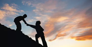 Silhouette image of two boys climbing a hill with one reaching out to help the other. 