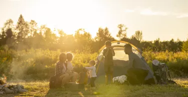 A small family settles into the campsite they have just finished setting up as they prepare to enjoy a vacation together. They are each dressed comfortably and some are finishing placing items in the tent while other enjoy sitting by the fire and relaxing