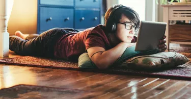 Teenager attending online classes at home during lockdown caused by COVID-19 world pandemic. He is looking at digital tablet with headset on his head while on the floor of his living room.