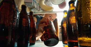 Caucasian woman takes cold refreshing beer from out the fridge, inside view from fridge of hand holding the bottle. Alcohole, domestic life, entertainment, drink concept. Cold cola or beer bottles.