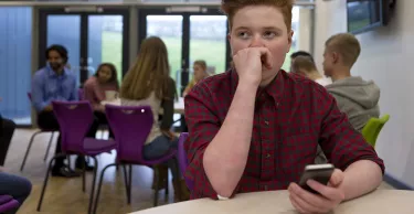 Stressed schoolboy sitting away from people at school. He has a smartphone in his hand and is looking away with his hand to his face.