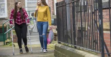 A young woman helps her friend who is on crutches by providing support and carrying her bag.