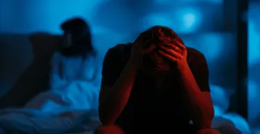 Depressed devastated man clutching head sitting on bed on distance from offended wife in dark red blue neon light on defocused background.