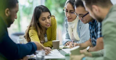A multi-ethnic group of business people are indoors in an office boardroom. They are sitting around a table and making plans.