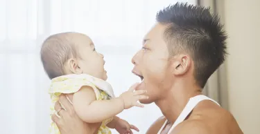 An adult holding a baby, with both facing each other indoors. The adult has their mouth open wide, and the baby is reaching toward their face.