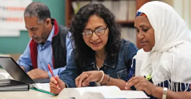 Three immigrant adults sit at a table studying; two women look at an open book together, while a man works on a laptop in the background.