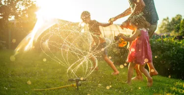 Father with children running through sprinkler at back yard in summer evening in sunset