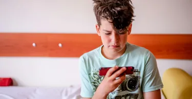 A young boy sits in his bedroom playing on a mobile device.