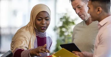 Mixed ethnic group of business professionals at a networking conference/meeting room. They are standing looking at digital tablets together. Muslim woman leads the business meeting.