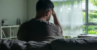 Young Asian man sitting on a sofa at home, looking sad and exhausted, struggling with depression and mental health issues.