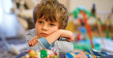 Image of a young boy sitting at a table with some colourful toys and he is resting his head on his arms. 