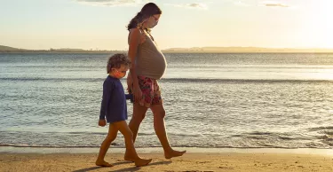 Pregnant mother exercising at the beach, holding her toddler's son hand by the sea, during sunset.