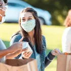 Two cheerful female friends fill paper bags with food donations as they volunteer during an outdoor food drive. They are wearing protective face masks as they are volunteering during the COVID-19 pandemic.