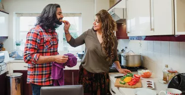 Mature woman is cooking a curry in the kitchen of her home. She is feeding her son a piece of sliced pepper as he does the dishes.