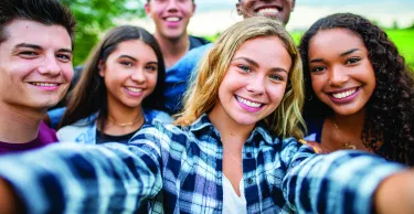 A multi-ethnic group of high school students are outdoors on a summer day. One girl is taking a selfie with the group.