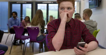 Stressed schoolboy sitting away from people at school. He has a smartphone in his hand and is looking away with his hand to his face.