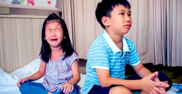 A photo of two children sitting on the floor. The boy who is older is sitting with his back to the younger girl and she is crying.
