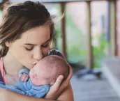 Young teenage woman is mother to newborn baby boy. She kisses him on the cheek as she holds him lovingly in her arms.