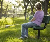 Rear view of one mature woman sitting on a park bench in the summer season. Bright sunny day with lush green grass and trees. The woman gazes off into the distance as she relaxes on a beautiful day. Solitude, lonliness, contemplation. She has short blond hair and wears a purple shirt and jeans. Copyspace to right in this tranquil nature scene.