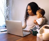 A young mixed race African American mother holds her daughter while taking notes at her dining table serving as a temporary remote work from home station with breast pump in foreground.