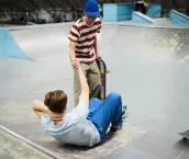 Two teenage boys are in a skate park. One boy is laying on the ground on his back, he has fallen. The other has extended his arm to help him back on his feet. 