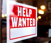 Sign text closeup for help wanted with red and white colors by entrance to store shop business building during corona virus covid 19 pandemic. Sign text closeup for help wanted with red and white colors by entrance to store shop business building during corona virus covid 19 pandemic.