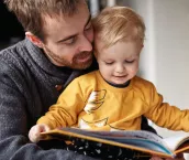 Cropped shot of an adorable smiling little boy reading a book while sitting with his father at home