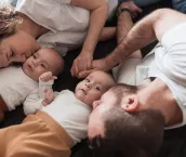 Happy young family with newborn twin babies lying on the bed together, parents holding hands and kissing their babies