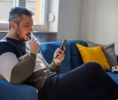 Man sitting on the sofa at home and using smart phone for home finances, smoking electronic cigarette and looking serious