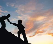 Silhouette image of two boys climbing a hill with one reaching out to help the other. 