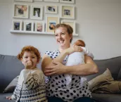 Mother with her two children in the living room of their home. The mother is holding a baby and the other little girl is laughing and looking at the camera while sucking her thumb.