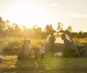 A small family settles into the campsite they have just finished setting up as they prepare to enjoy a vacation together. They are each dressed comfortably and some are finishing placing items in the tent while other enjoy sitting by the fire and relaxing
