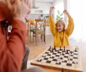 A delighted granddaughter raises her arms in celebration after winning a game of chess at home. Her face lights up with joy and pride as she revels in her success, while the peaceful home setting adds to the warmth of this special moment of achievement.