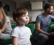 Upset frustrated little boy tired of parents fight, toddler son looking out window reaming that family conflicts would stop while parents sitting separately on sofa, suffering from mom and dad quarrel