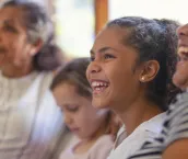 Multi generational women on the sofa. The young girls are sisters. One is a teenager on is younger. There is also a mother and grandmother. They are all happy and smiling. They are all Australian Aboriginals.