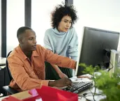 Waist-up view of casually dressed Black and Hispanic associates collaborating as they sit and stand at flexible workstation, looking at desktop PC monitor.