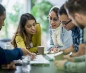 A multi-ethnic group of business people are indoors in an office boardroom. They are sitting around a table and making plans.