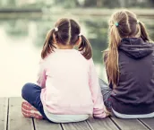 Two girls sitting on a wooden pier, their backs to the lens, surrounded by a serene waterbody.
