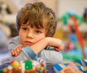 Image of a young boy sitting at a table with some colourful toys and he is resting his head on his arms. 